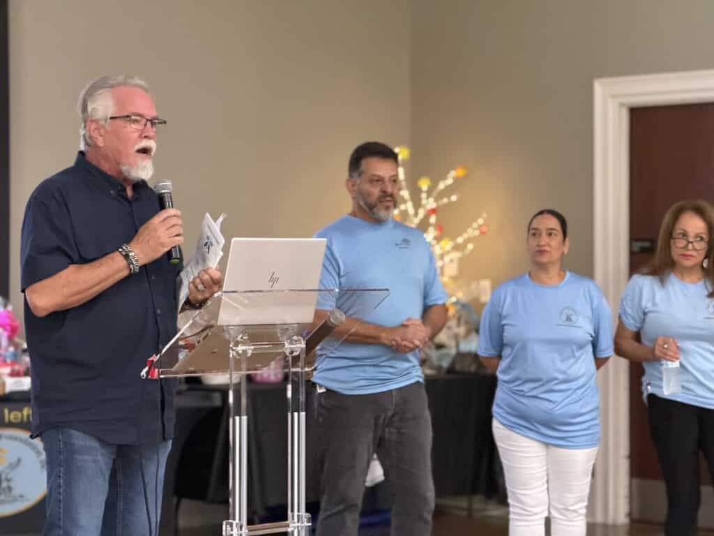Man at a podium speaking beside volunteers in light blue shirts