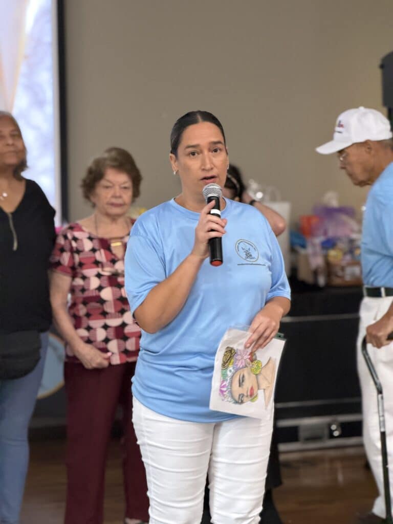 Woman in a light blue shirt speaking into a microphone to guests